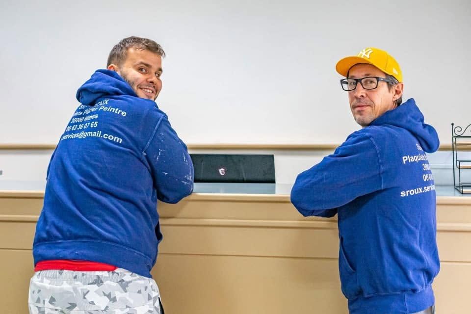 Deux artisans en sweat bleu floqué « S. Roux, plaquiste, plâtrier, peintre, Nîmes » sur un chantier.